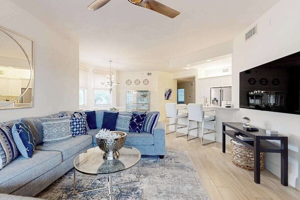 A modern living room with a sectional sofa, a coffee table, and a dining area with a TV and kitchen appliances. - Luxurious condo with on-site pool, private extended balcony, & W/D (Naples)
