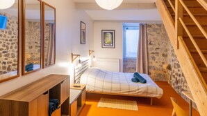 Quarto com cama, escrivaninha de madeira, espelhos, paredes de pedra, escada e janela.