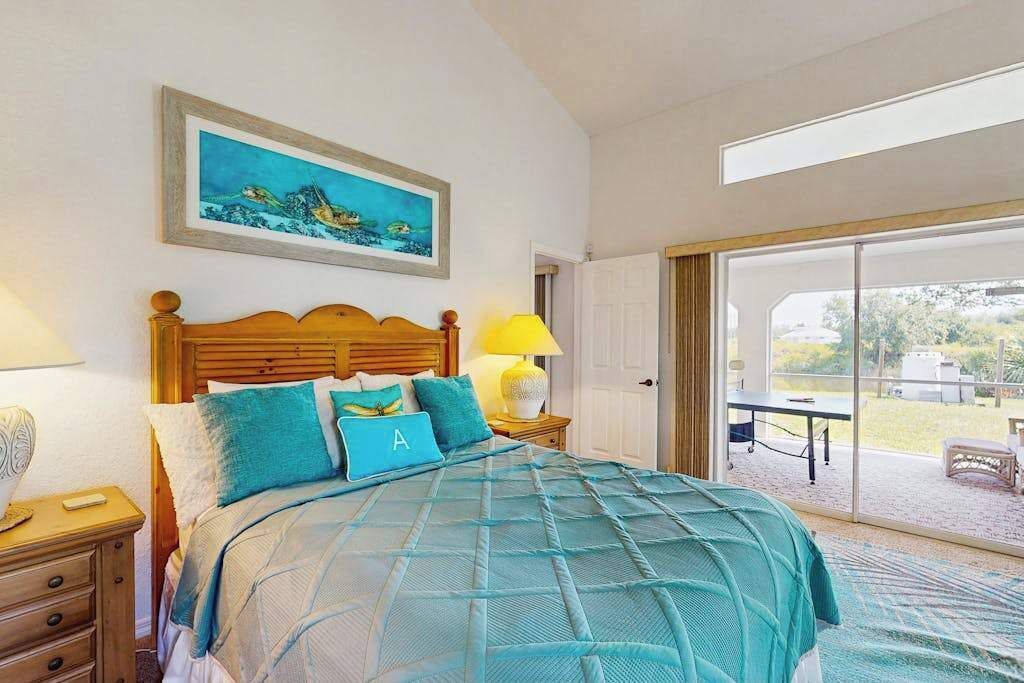 A bedroom with a bed, a nightstand, a lamp, a painting, and a sliding glass door leading to an outdoor area.