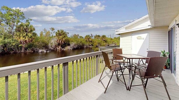 A deck with a table and chairs overlooking a river, surrounded by greenery and a building with a garage. - Pool, Game Room & Waterfront | 30+ Days (Port Charlotte)