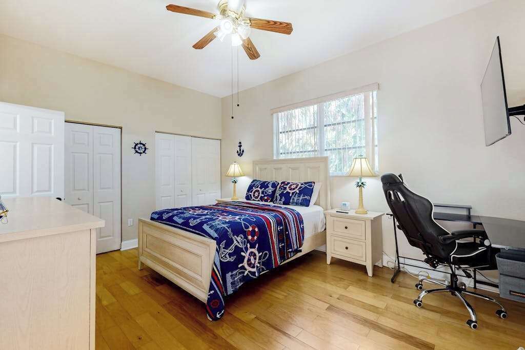 A bedroom with a bed, bedside tables, a desk, a chair, a ceiling fan, and a window.
