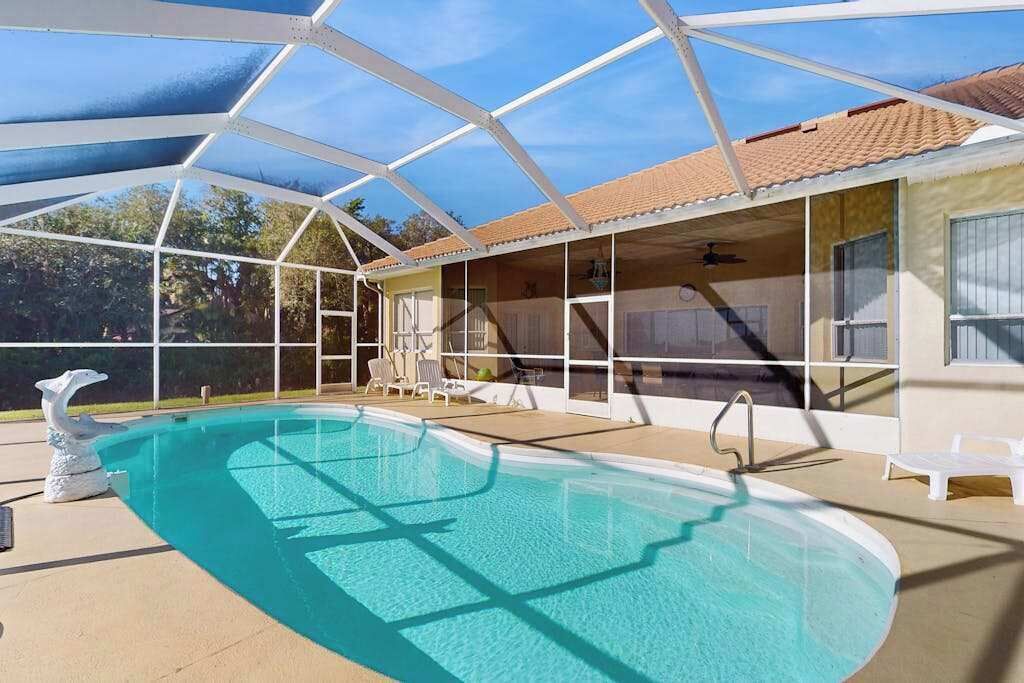 A swimming pool with a glass enclosure, white chairs, and a covered patio area.