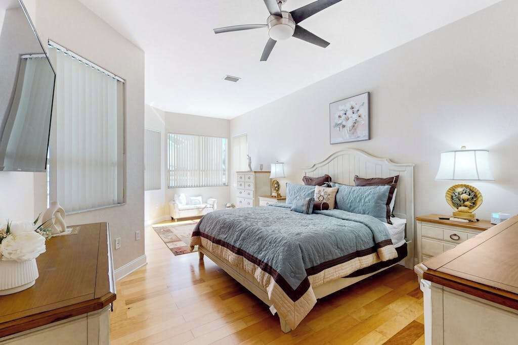 Une chambre à coucher comprenant un lit, des tables de chevet, un ventilateur de plafond et une fenêtre avec des stores.