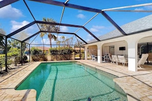 A covered swimming pool with a geometric roof structure, surrounded by a patio area with lounge chairs and a view of palm trees. - Sunny Snowbird Retreat with a Pool, Furnished Lanai, & Prime Corner Lot (Port Charlotte)