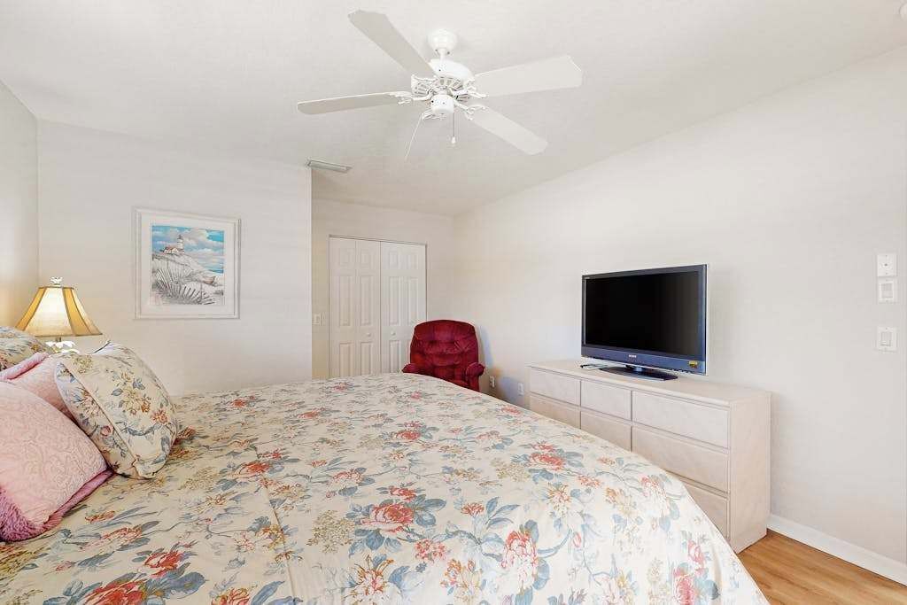 Une chambre à coucher avec un lit, une chaise rouge, un téléviseur posé sur une commode, un ventilateur de plafond et une porte.