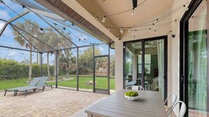 Une terrasse couverte avec une table et des chaises, une porte vitrée donnant sur une pièce, et une vue sur une pelouse et des arbres.