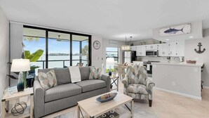 A modern living room with a sofa, armchair, and coffee table, a kitchen with white cabinets, and a large window offering a view. - Water view condo with shared pool, panoramic balcony views, and WIFI (Marco Island)