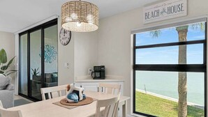 A dining area with a table, chairs, a woven light fixture, a clock, and a view of the beach. - Water view condo with shared pool, panoramic balcony views, and WIFI (Marco Island)