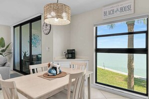A dining area with a table, chairs, a woven light fixture, a clock, and a view of the beach. - Water view condo with shared pool, panoramic balcony views, and WIFI (Marco Island)