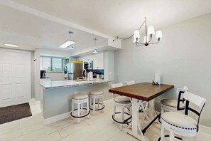 A modern kitchen with a central island, white bar stools, and a dining table with chairs. - Beachfront condo with screened balcony, community pool, and internet (Fort Myers Beach)