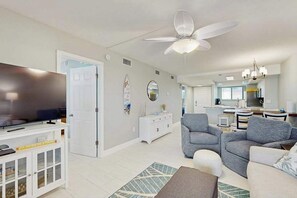 A modern living room with a flat-screen TV, grey sofas, a ceiling fan, and a kitchen in the background. - Beachfront condo with screened balcony, community pool, and internet (Fort Myers Beach)