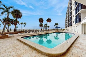 A swimming pool with lounge chairs, palm trees, and a building with balconies. - Beachfront condo with screened balcony, community pool, and internet (Fort Myers Beach)