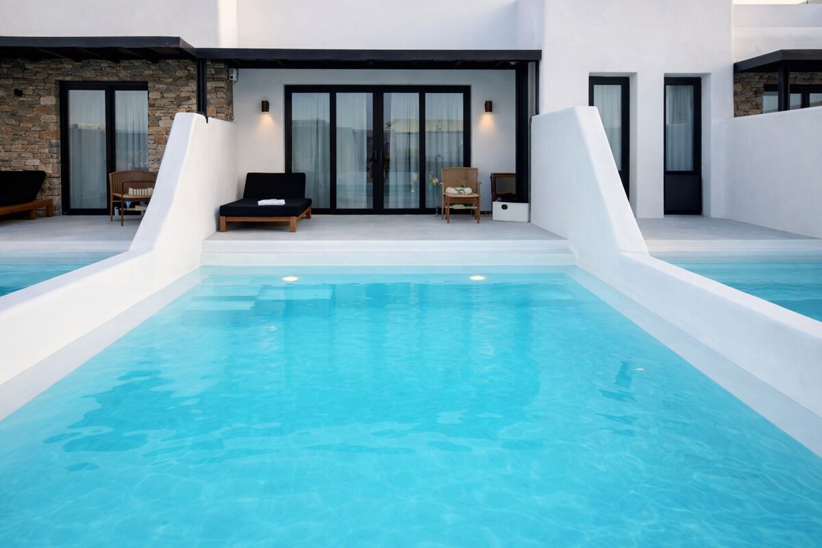 Un área de piscina moderna con una piscina azul cristalina, dos sillas de descanso y un muro de piedra.