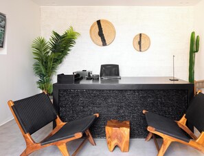A reception desk with a black textured counter, two wooden chairs with black mesh seats, a potted plant, and two wall-mounted wooden coasters. - Hotiday Suites Mykonos (Mykonos)