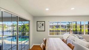 Un espace repas comprenant une table et des chaises en bois, un canapé et de grandes fenêtres donnant sur une piscine et un espace verdoyant.