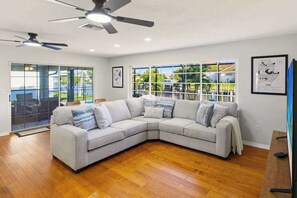 Ein modernes Wohnzimmer mit Ecksofa, Holzboden, Deckenventilatoren, großen Fenstern und Blick ins Freie.