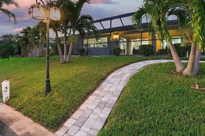 A pathway leading to a modern building with glass windows, surrounded by well-maintained grass and palm trees. - Waterfront home, with private pool and lanai, Gulf access with fishing dock (Punta Gorda)