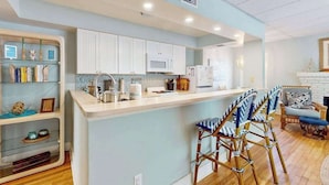 A modern kitchen with white cabinets, a central island, bar stools, and a dining area with a wicker chair. - Dog-friendly duplex with outdoor living - walk to beach & downtown (Venice)