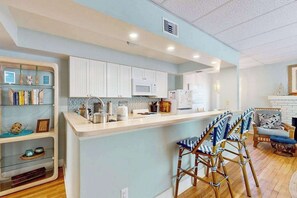 A modern kitchen with white cabinets, a central island, bar stools, and a dining area with a wicker chair. - Dog-friendly duplex with outdoor living - walk to beach & downtown (Venice)