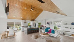 Una cocina moderna con una isla central, un comedor con mesa y sillas de madera, una sala de estar con sofá blanco y cojines coloridos, y un techo con vigas de madera e iluminación empotrada.
