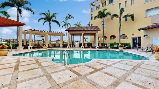 Piscina com espreguiçadeiras e área de estar coberta, cercada por palmeiras e um edifício de vários andares.