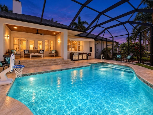 A swimming pool with a sunken seating area, a covered patio with outdoor furniture, and a view of the surrounding landscape.