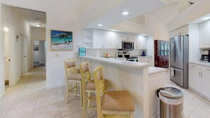 A kitchen with white cabinets, a stainless steel refrigerator, and a bar area with chairs. - Canal-front home with private pool, pool spa, dock, boat access, grill, gym, W/D (Marco Island)