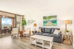 Ein modernes Wohnzimmer mit grauem Sofa, weißem Couchtisch, Korbstühlen und Blick auf den Ozean durch große Fenster.