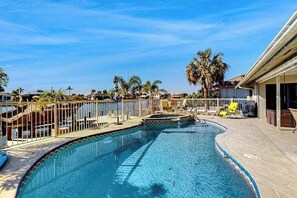 Ein Swimmingpool mit umzäunter Terrasse, Palmen und einem Gebäude mit überdachtem Bereich.