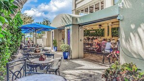 Uma área de pátio ao ar livre com mesas, cadeiras, plantas em vasos e entrada de restaurante.