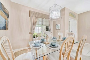 A dining room with a glass table, white chairs, a chandelier, and a painting on the wall.