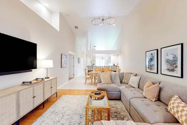 A modern living room with a flat-screen TV, a sofa, a coffee table, and a dining area in the background. - 2nd-floor lakefront condo with heated pool, lake view, balcony, bikes, fast WiFi (Naples)