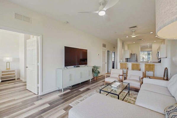 A modern living room with a flat-screen TV, a sofa, a coffee table, and a dining area with chairs and a kitchen in the background. - Newly Remodeled Chic Condo on the GreenLinks golf course with lanai, pool. (Naples)