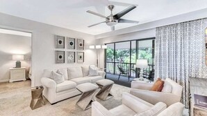 Ein modernes Wohnzimmer mit Deckenventilator, einer Sofagarnitur, einem Couchtisch und Blick durch große Fenster.