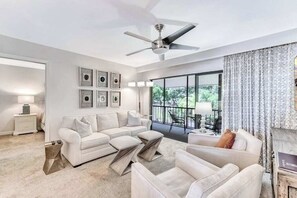 Ein modernes Wohnzimmer mit Deckenventilator, einer Sofagarnitur, einem Couchtisch und Blick durch große Fenster.