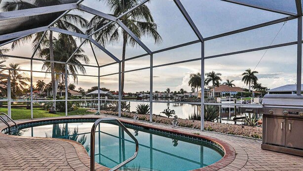 Piscina com vista para um lago e palmeiras, rodeada por um pátio de tijolos e uma área coberta com churrasqueira.