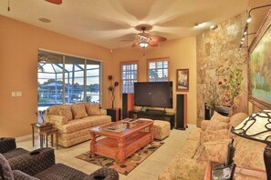 A living room with a ceiling fan, a flat-screen TV, sofas, a coffee table, and a fireplace. - Upscale waterfront home with private dock, saltwater pool, lanai, grill, & W/D (Naples)