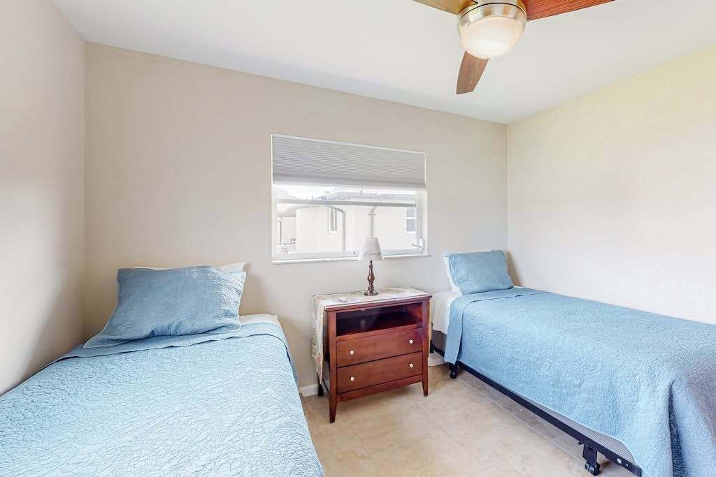 Une chambre avec deux lits, une table de chevet, un ventilateur de plafond et une fenêtre avec des stores.