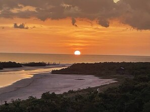 A sunset over a coastal landscape with a body of water, vegetation, and a distant structure. - Water view condo w/ access to community amenities, wifi, & outdoor private patio (Naples)