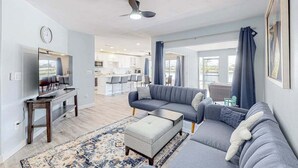 Un salon moderne comprenant un canapé, un pouf et un téléviseur, et donnant sur un espace cuisine avec un coin repas.