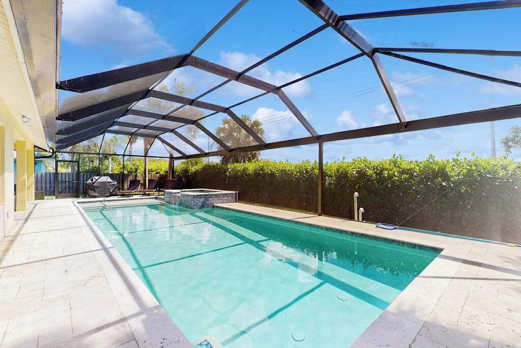 En swimmingpool med et glasafskærmning, omgivet af en fliseterrasse og en hæk.