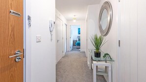 Un pasillo con una puerta de madera, una pared blanca, una mesa de vidrio y una planta en maceta, y un espejo.