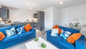 Una sala moderna con dos sofás azules, una mesa de centro de vidrio y una cocina al fondo.