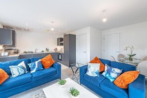 A modern living room with two blue sofas, a glass coffee table, and a kitchen in the background.