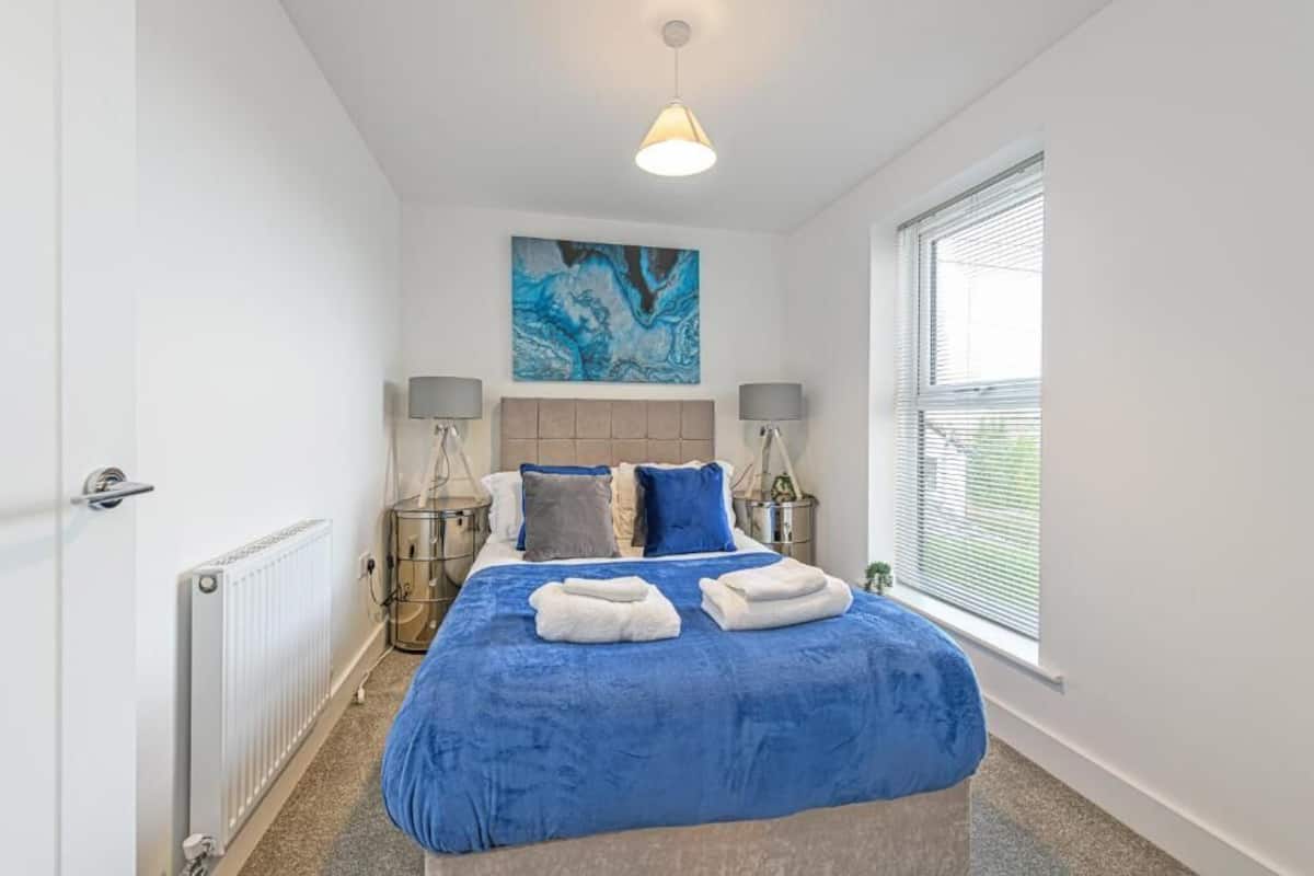 A bedroom with a large bed, two bedside tables, a radiator, a window with blinds, and a ceiling light.
