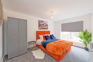 Camera da letto con un letto, un armadio, un tavolino, un tappeto bianco, una finestra con persiane e una pianta.