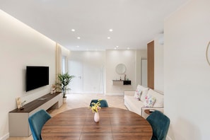 A modern living room with a round wooden dining table, a flat-screen TV mounted on the wall, a sofa with floral cushions, and a potted plant.