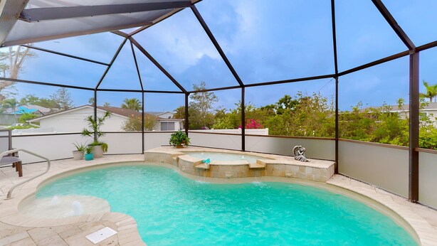 A swimming pool with a curved design, surrounded by a covered area with a metal frame, potted plants, and a view of neighboring houses. - Canal-Front w/Private Pool & Dock – Your Florida Escape Starts Here (Punta Gorda)
