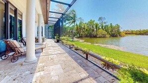A covered outdoor area with a view of a lake, stone flooring, and lounge chairs. - Deluxe home in a resort community with amazing views, pool, & hot tub (Naples)