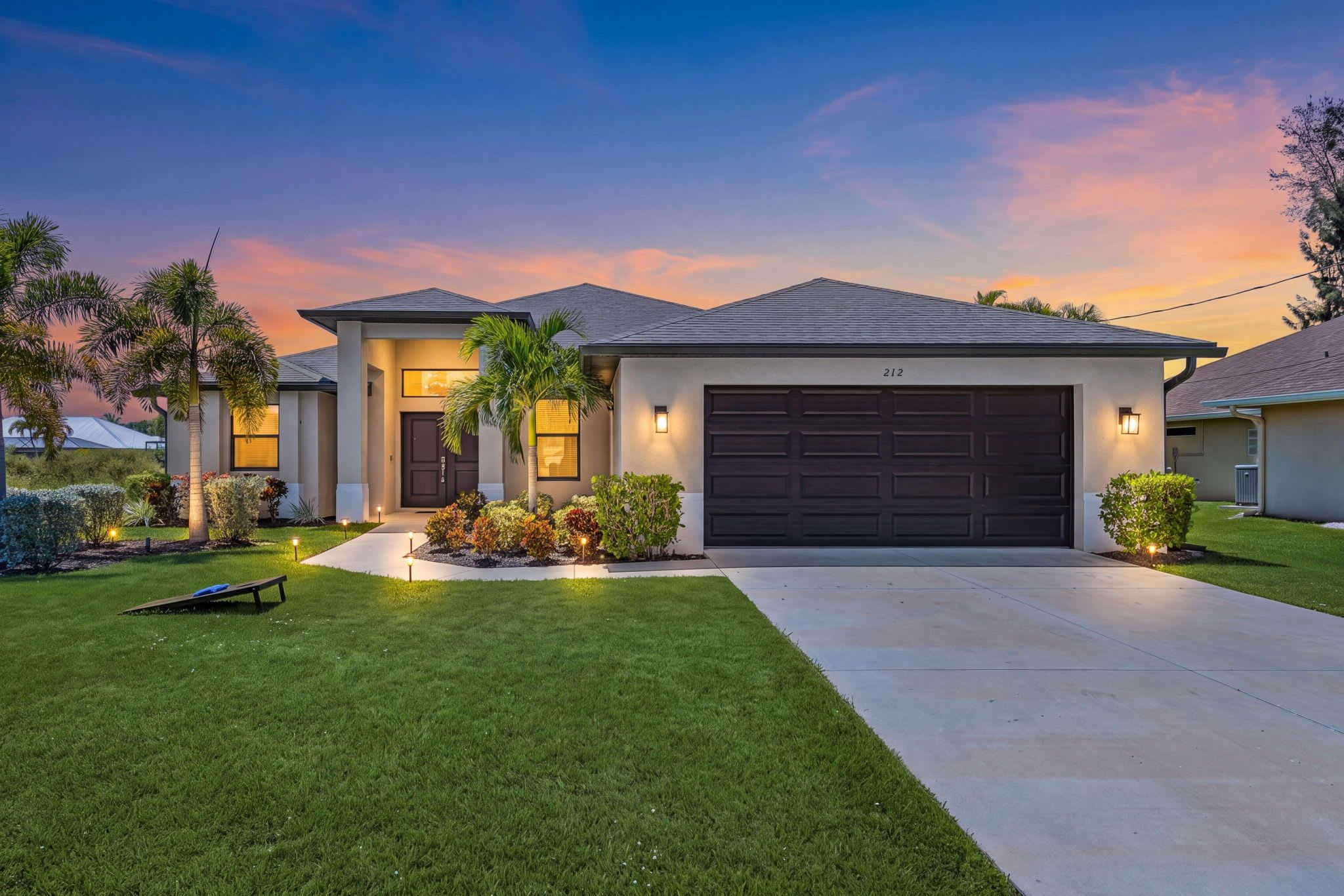 A modern two-story house with a dark garage door, well-maintained lawn, and a variety of shrubs and plants.